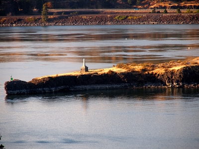 [A skinny finger of an island in the river with a two-tier grey monument on it.]